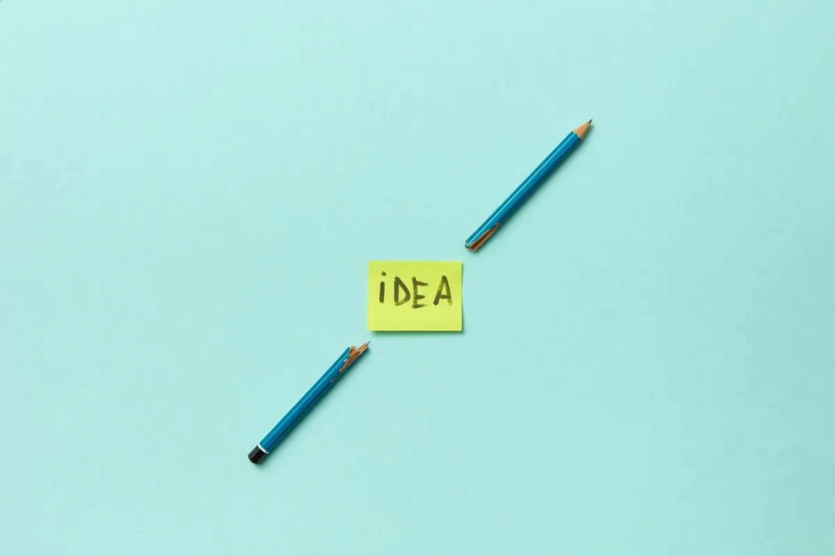 Un lápiz partido en dos junto a una nota adhesiva amarilla con la palabra idea, sobre fondo azul claro
