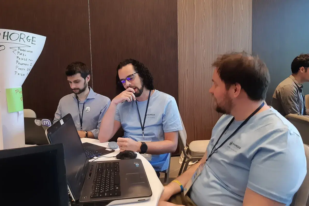 Tres participantes conversan alrededor de una mesa con un portátil durante el Wikimedia Hackathon 2024